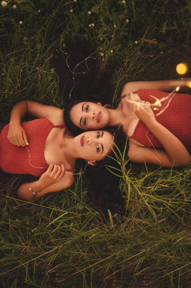 Two sisters lay on the grass with their heads together, holding a string of lilghts.