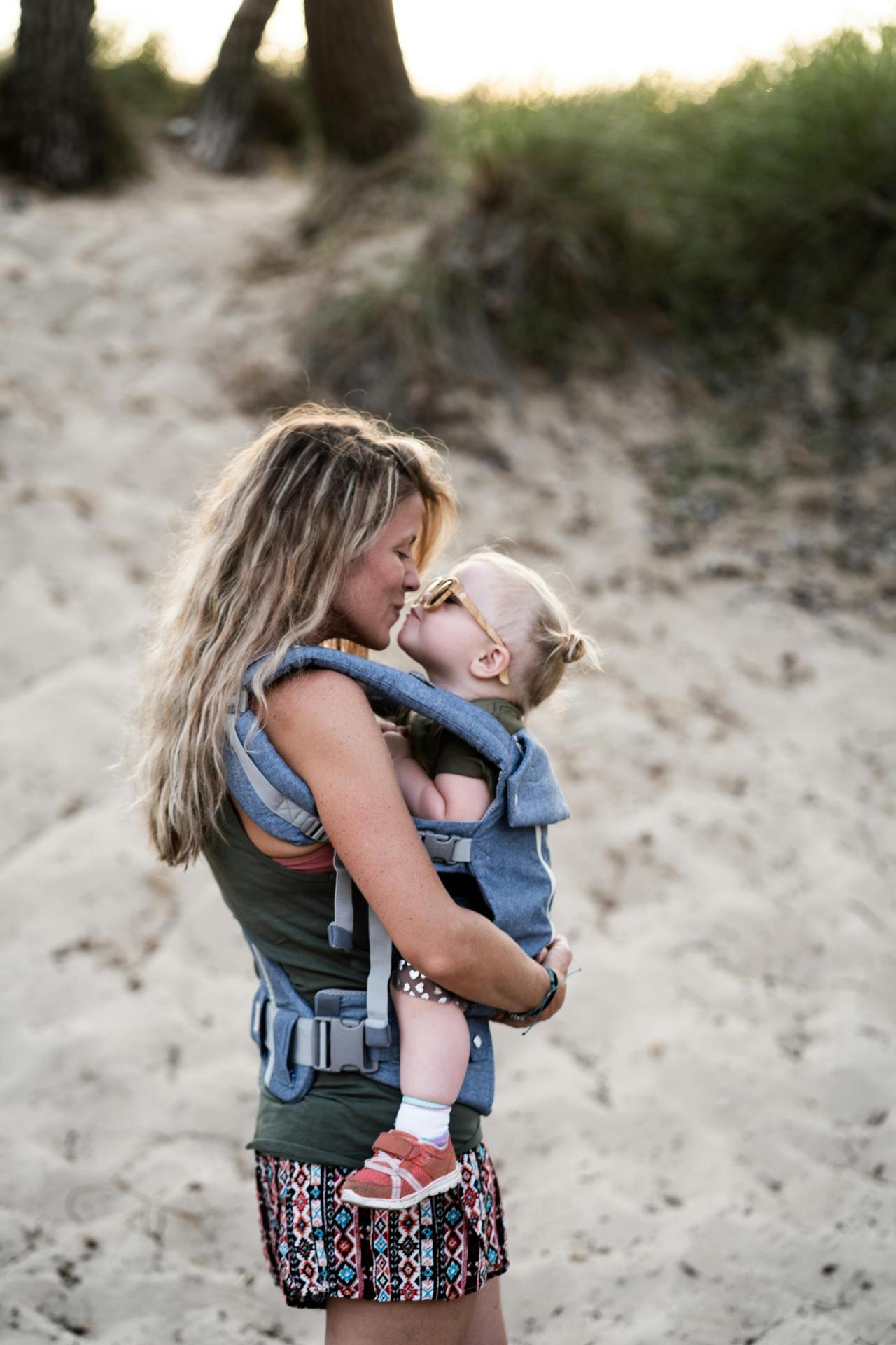 A mother holds her daughter in a sling and they face each other, smiling.