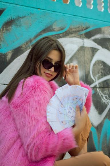 A woman in a pink fur coat and sunglasses fans several money bills.