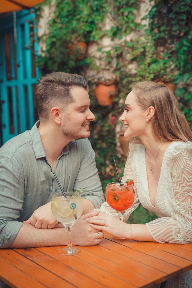 A man and woman sit together at a table, smiling and holding hands with two drinks in front of them.