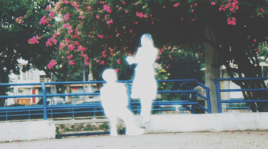 Two heavily overexposed white silhouettes of a child and an adult standing near a flowering tree with pink blossoms and blue railings in a park.