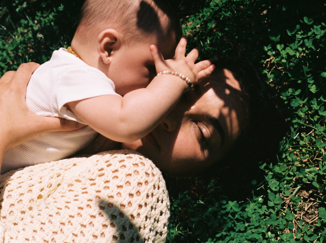 Mother lying in a clover field holding her baby, who reaches a small hand toward her face.