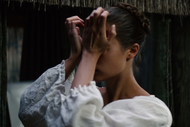Woman in a white puff-sleeve dress covering her face with both hands, head bowed, beside a thatched structure.
