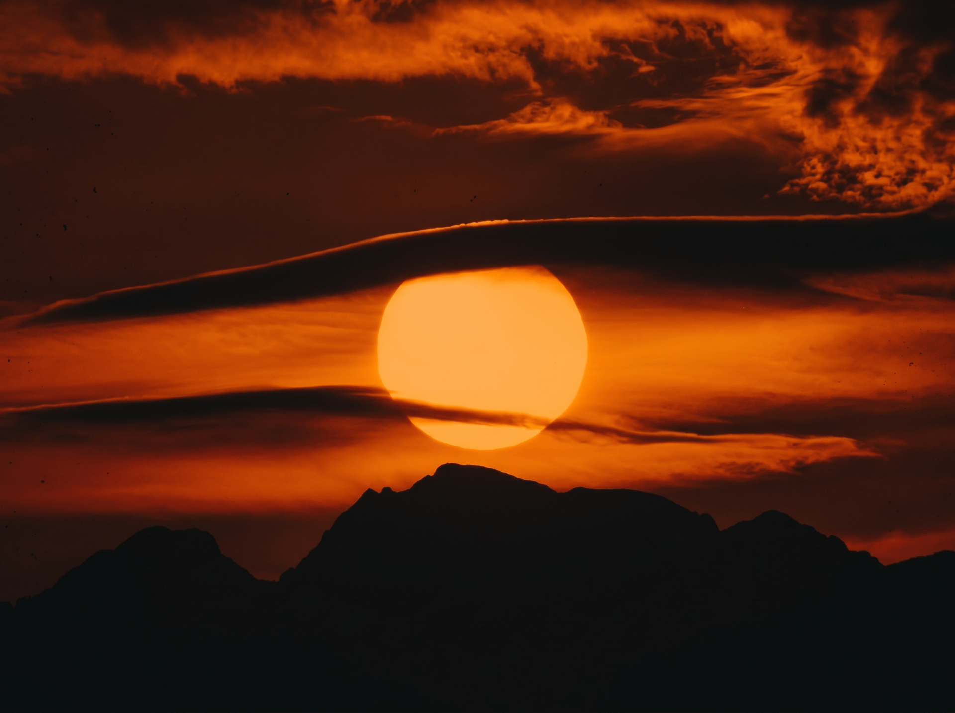 Large orange sun setting behind a dark mountain silhouette against a deep red and orange cloudy sky.