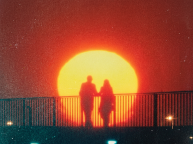 Silhouettes of two people holding hands on a bridge railing against a large glowing sun and deep red sky.
