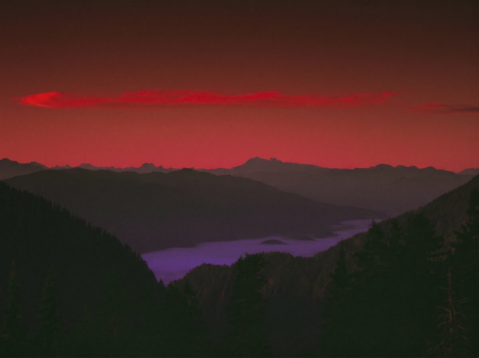 Deep red and crimson sunset sky over silhouetted mountain ridges with a purple-hued lake nestled in a forested valley below.