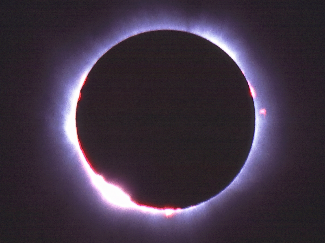 Total solar eclipse showing the moon's dark silhouette encircled by a white corona and red solar prominences against a dark sky.