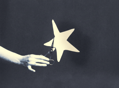 A hand holding scissors cutting out a large paper star against a dark background, in a moody blue-toned monochrome style.