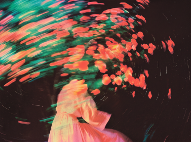 Motion-blurred figure in a pink dress surrounded by swirling orange flowers and green leaves against a dark background.
