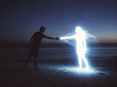 Silhouetted man reaching toward a glowing, ghost-like female figure on a beach at night under a starry sky.
