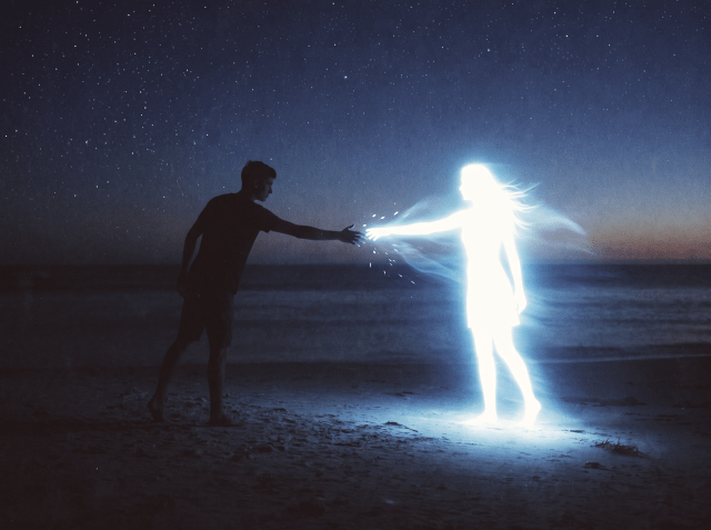 A man's silhouette reaches out toward a glowing, ghost-like female figure on a dark beach under a starry night sky.