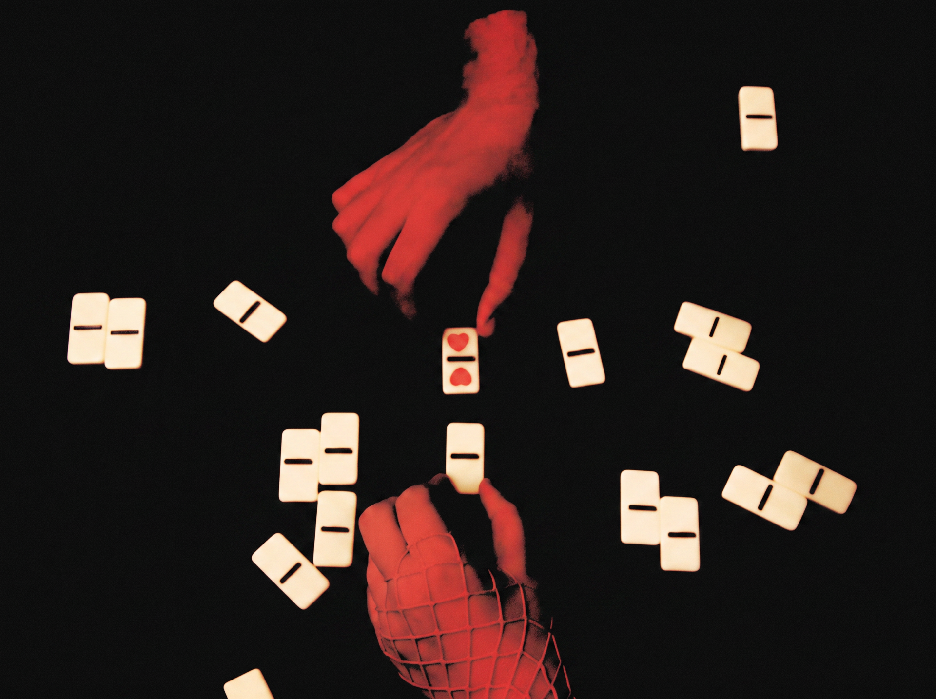 Two hands bathed in red light reaching toward scattered domino tiles against a black background, with one domino showing red heart symbols.