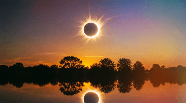 Total solar eclipse with radiating corona over a calm lake at sunset, silhouetted trees lining the horizon and the eclipse reflected in the water