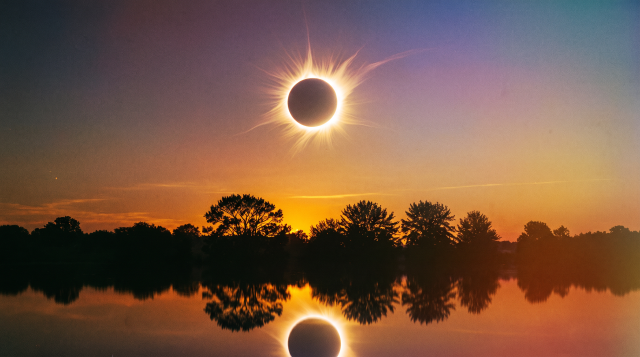 Total solar eclipse with radiating corona over a calm lake at sunset, silhouetted trees lining the horizon and the eclipse reflected in the water