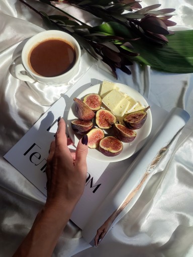 Overhead view of a hand with dark nail polish reaching toward a white plate of halved figs and sliced cheese next to a cup of black coffee on white fabric.