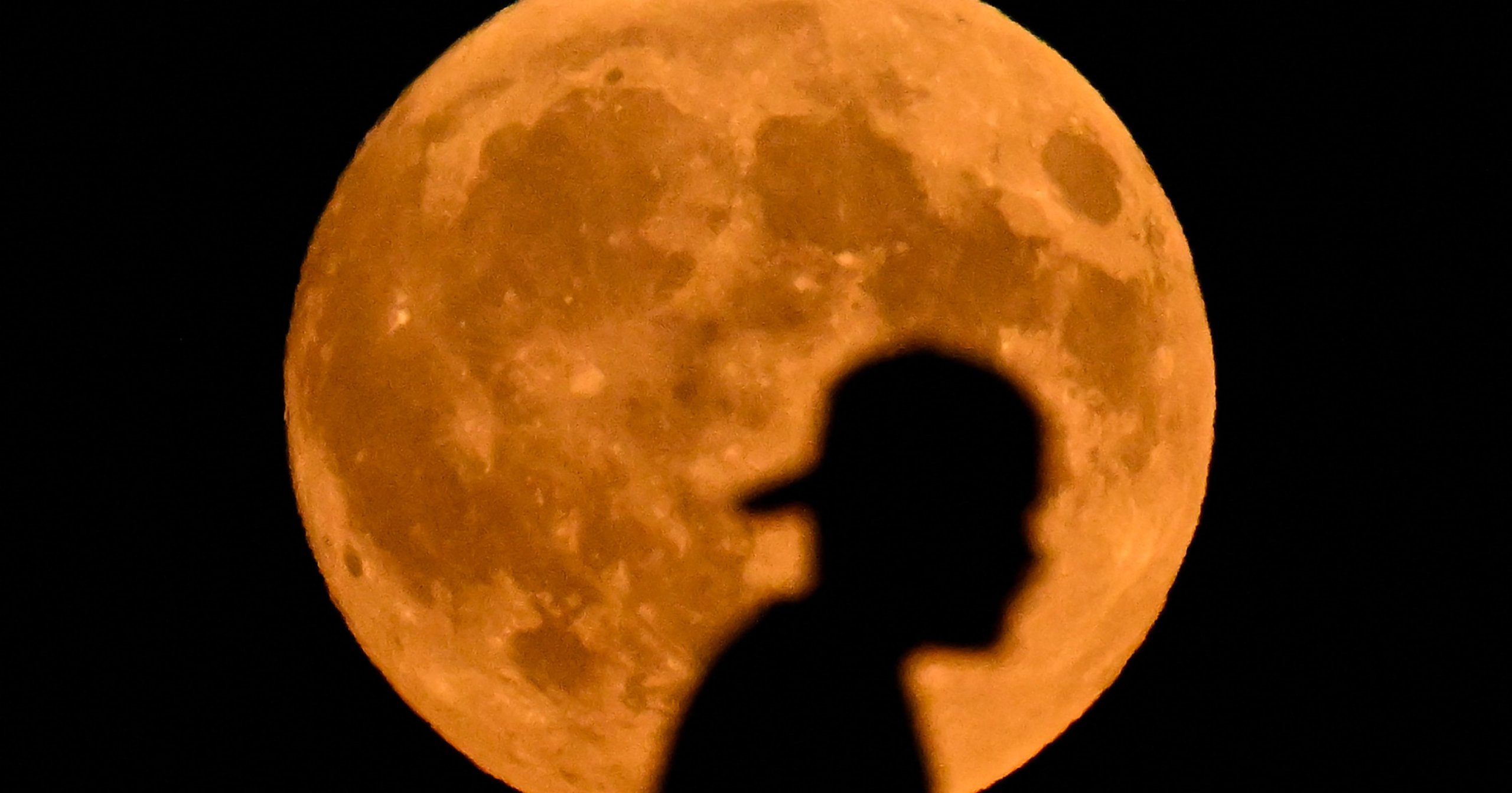Silhouette of a person crossing a bridge against a large, orange full moon in a dark night sky.