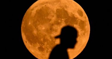 Silhouette of a person crossing a bridge against a large, orange full moon in a dark night sky.