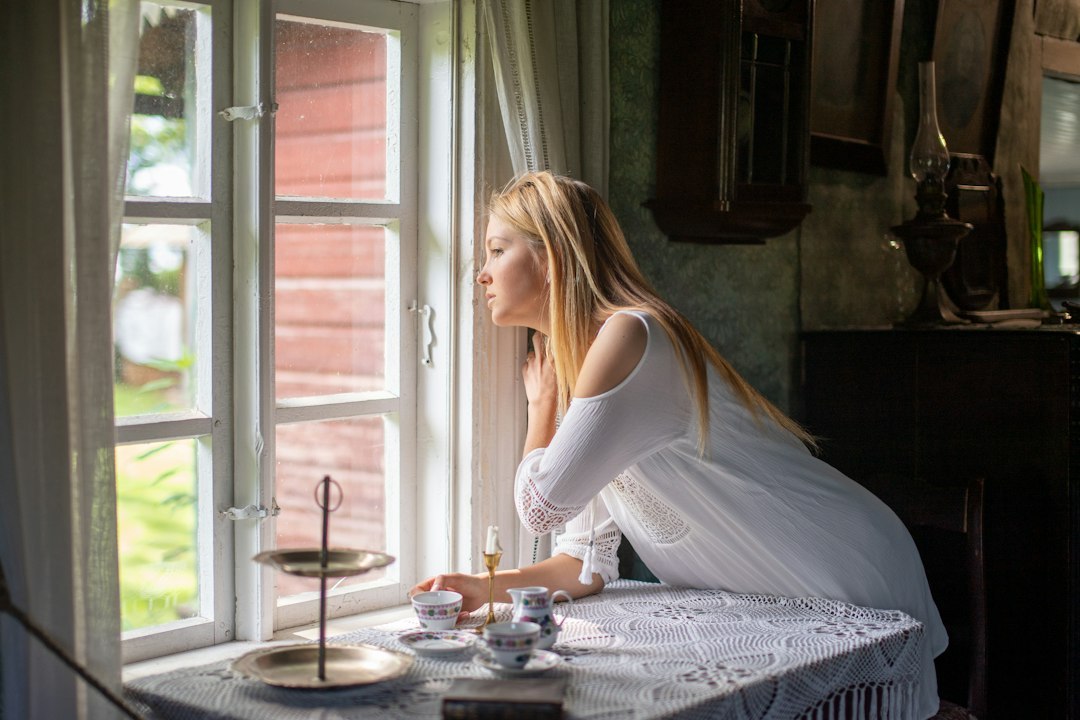 Thoughtful blonde woman in a white dress looking out the window of a rustic vintage cottage during morning tea.