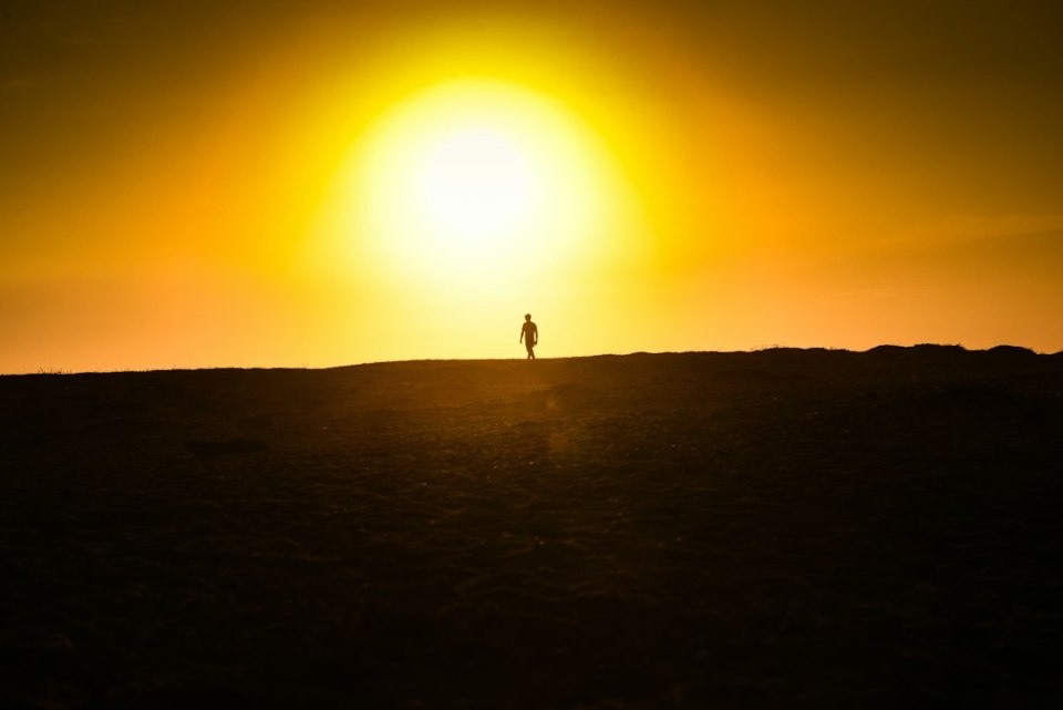 A lone person in silhouette walks along a ridge with a bright sun and orange sky behind them
