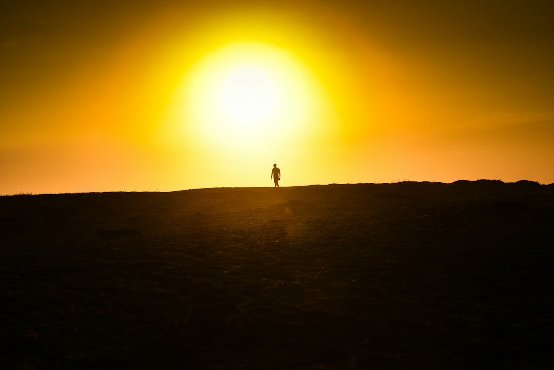 A lone person in silhouette walks along a ridge with a bright sun and orange sky behind them