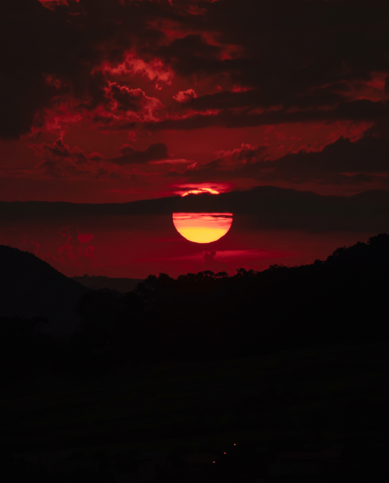 A red-orange sun setting behind clouds above dark hills.