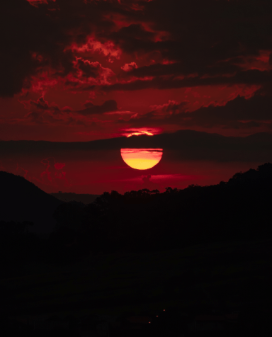 A red-orange sun setting behind clouds above dark hills.