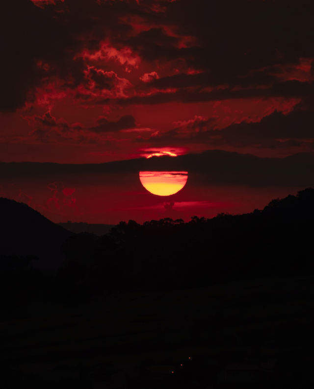A red-orange sun setting behind clouds above dark hills.