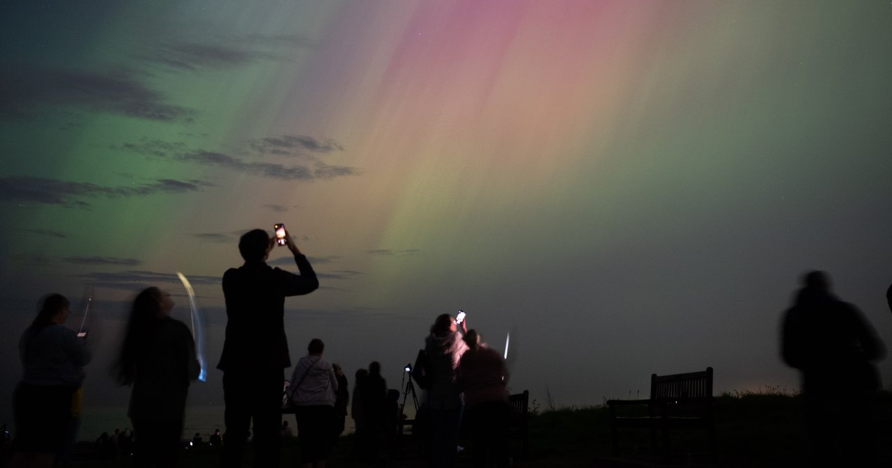 Silhouetted crowd holding up phones to photograph green and pink northern lights glowing over the sea near a lighthouse at night.